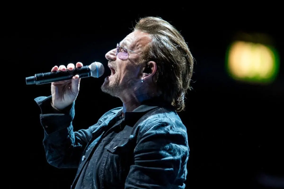 Bono Hewson sings passionately into a microphone onstage, wearing tinted glasses and a dark jacket against a black background.