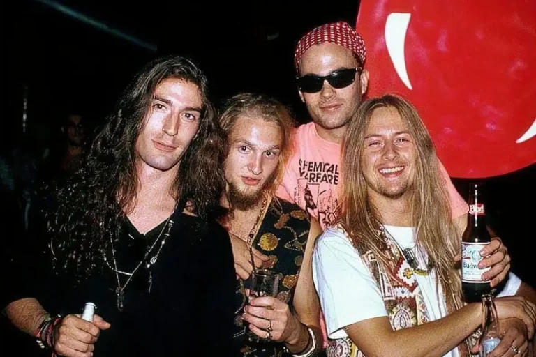 Alice in Chains band stand closely together backstage, smiling and holding drinks under dim concert lighting.