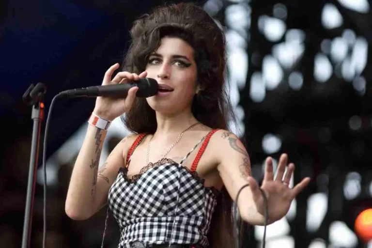 Amy Winehouse singing into a microphone on stage, wearing a checkered dress with her signature beehive hairstyle.