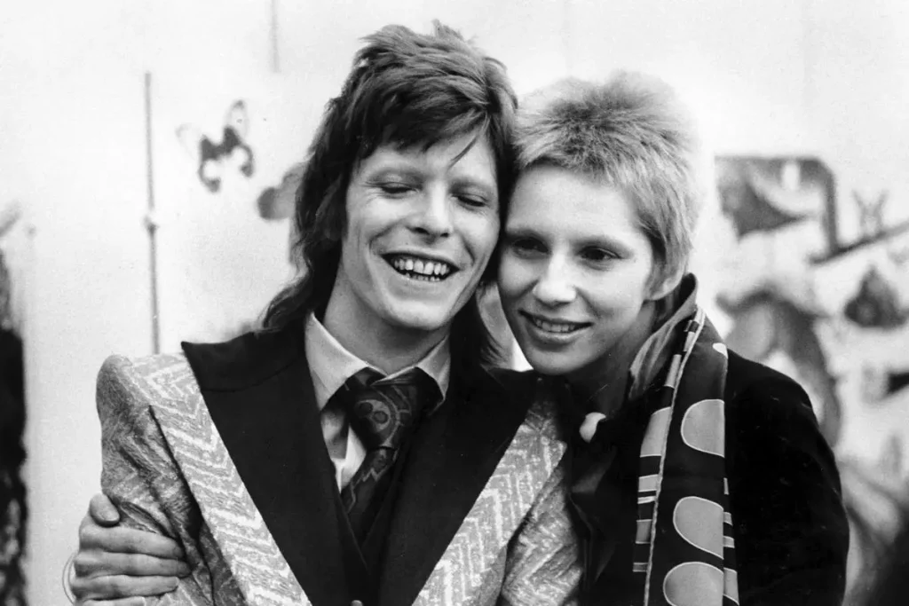 Angie and David Bowie stands close together, with the man wearing a patterned suit jacket and the woman leaning affectionately against him as they pose for the camera.