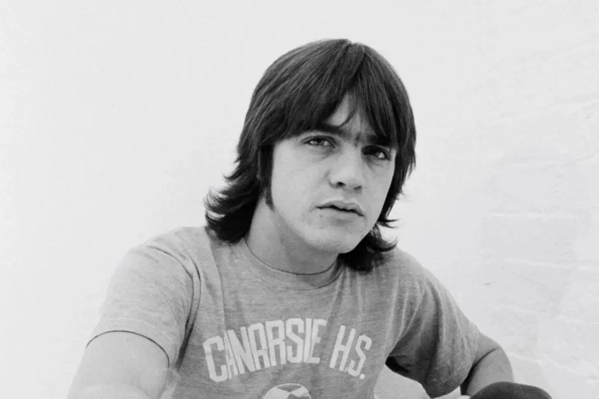 Angus Young with shaggy hair looks directly at the camera while wearing a T-shirt that reads “Canarsie H.S.” against a plain white background.