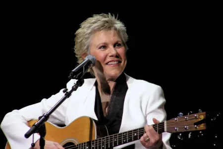 Anne Murray smiling while playing an acoustic guitar on stage.