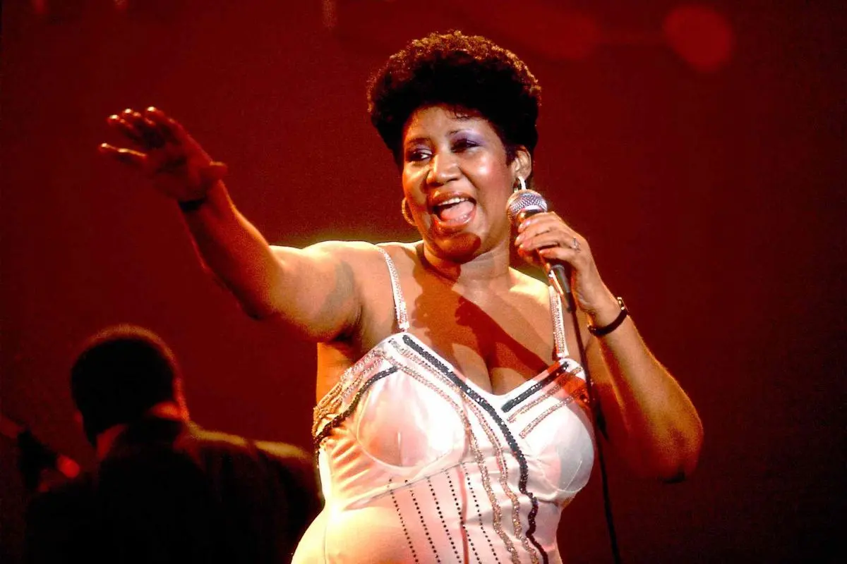 Aretha Franklin performing live on stage, singing into a microphone under red stage lights.