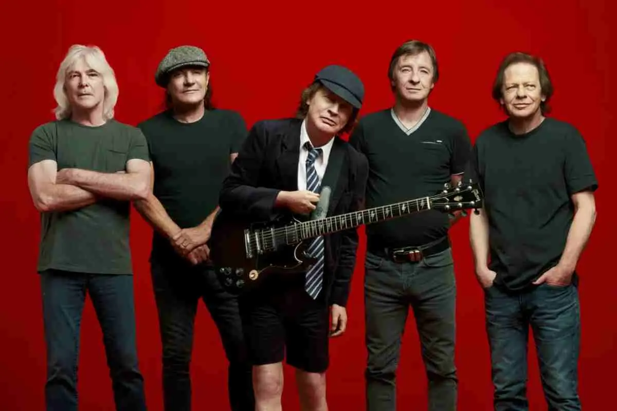 AC/DC posing as a group against a red background, with Angus Young standing front and center holding a guitar and wearing his signature schoolboy-style outfit.