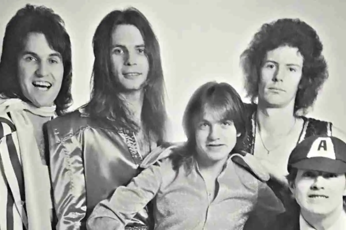 Early-era AC/DC band members posing together in a black-and-white promotional photo, wearing 1970s rock fashion with long hair and casual stage attire.