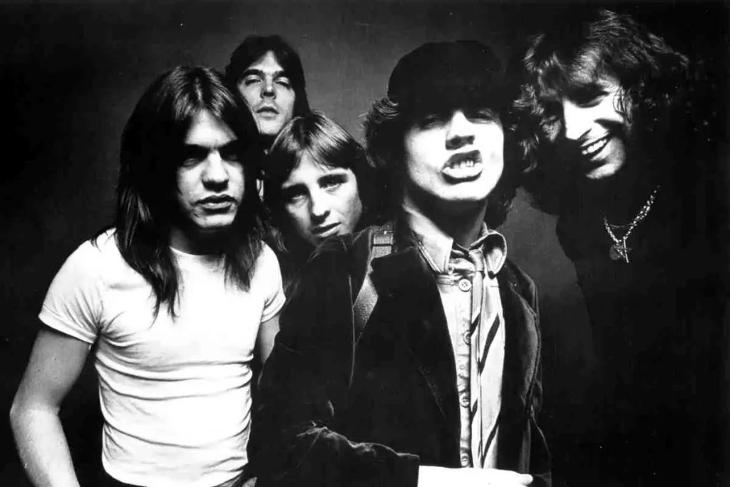 AC/DC band members posing closely together in a black-and-white portrait, featuring Angus Young in a cap at the front with bandmates behind him, all wearing classic rock-era clothing.