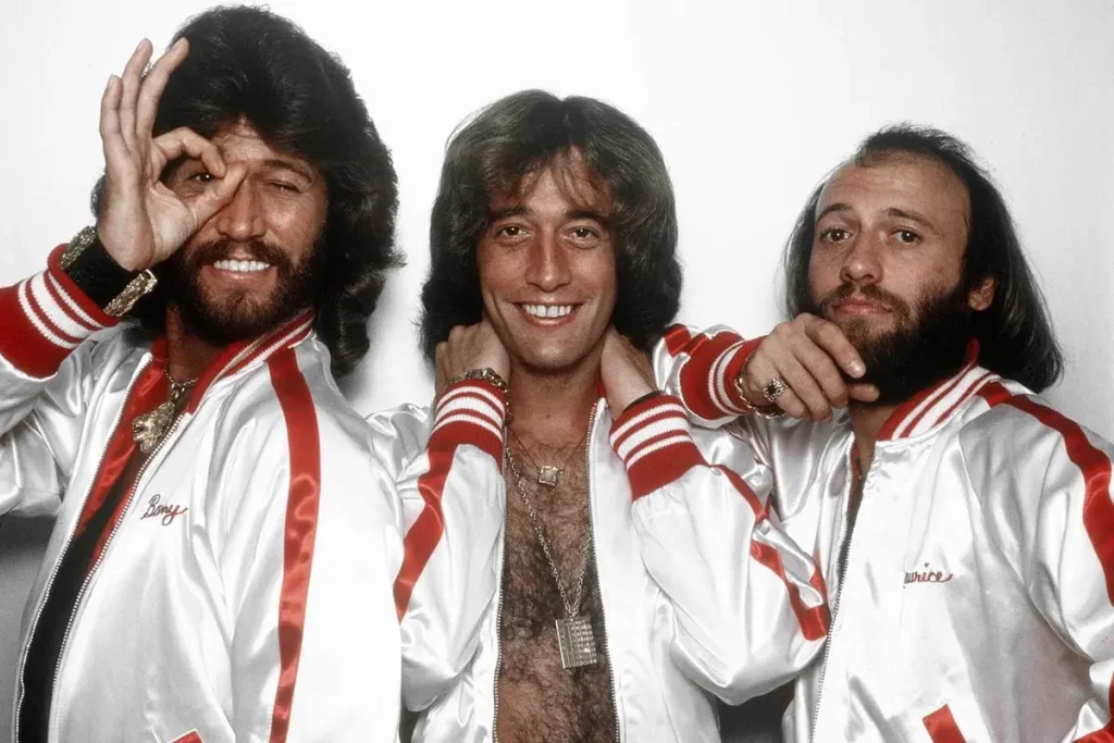 Bee Gees pose together in matching white satin jackets with red stripes.