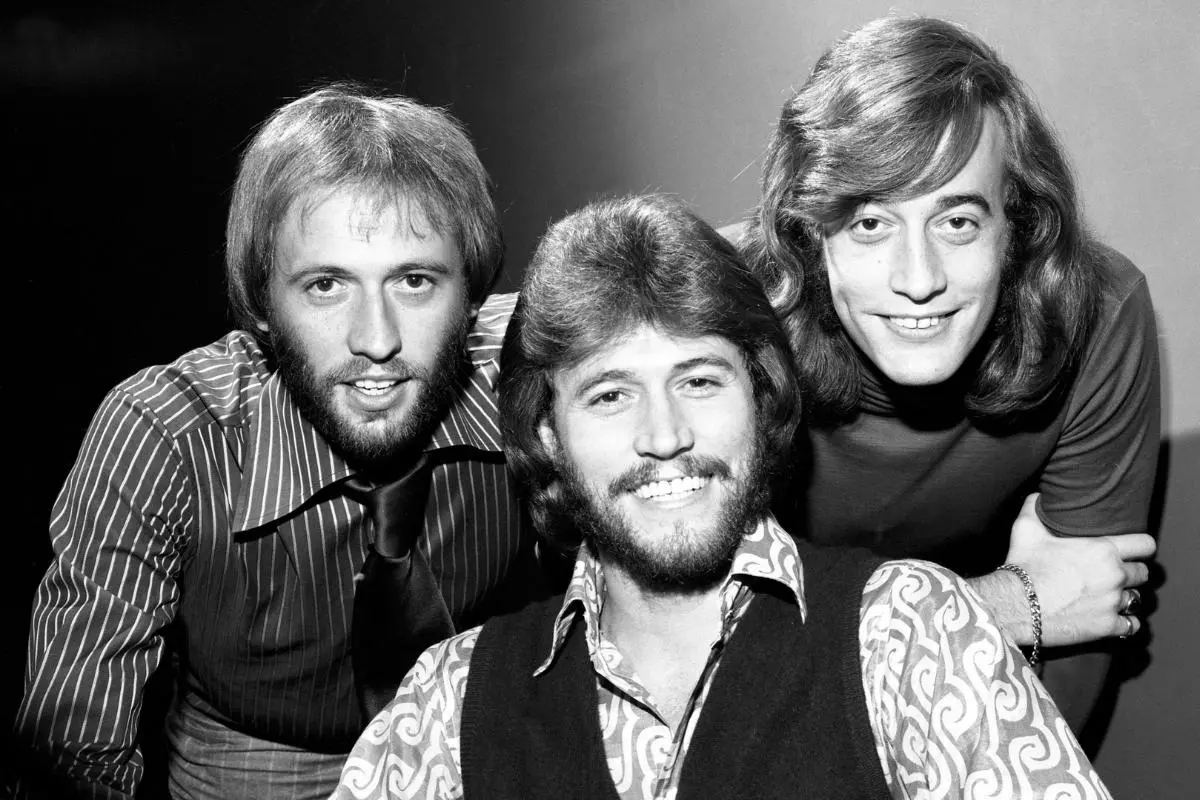 Bee Gees leaning close together, smiling at the camera, wearing 1970s-style shirts and vests.
