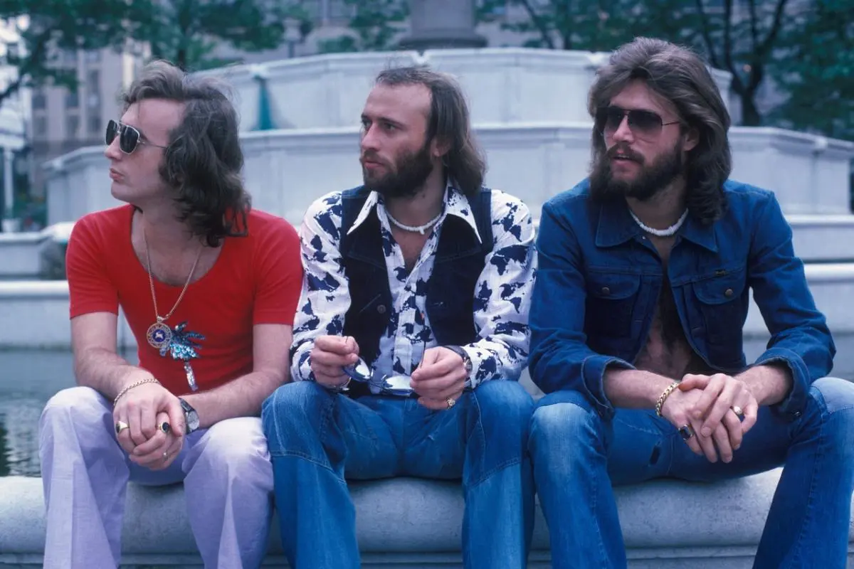 Bee Gees sit side by side on a stone ledge outdoors.