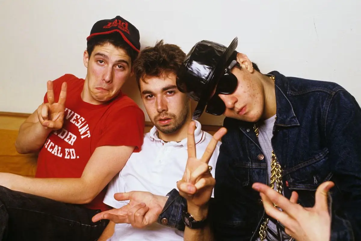 Beastie Boys  pose indoors making peace signs and exaggerated expressions, dressed casually with hats and layered clothing.
