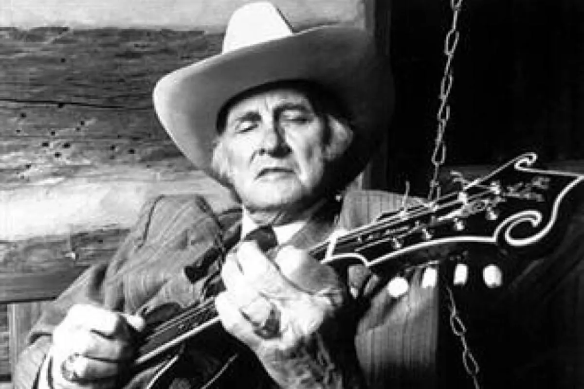 Bill Monroe wearing a cowboy hat sits on a wooden porch, playing a mandolin with a calm, reflective expression.