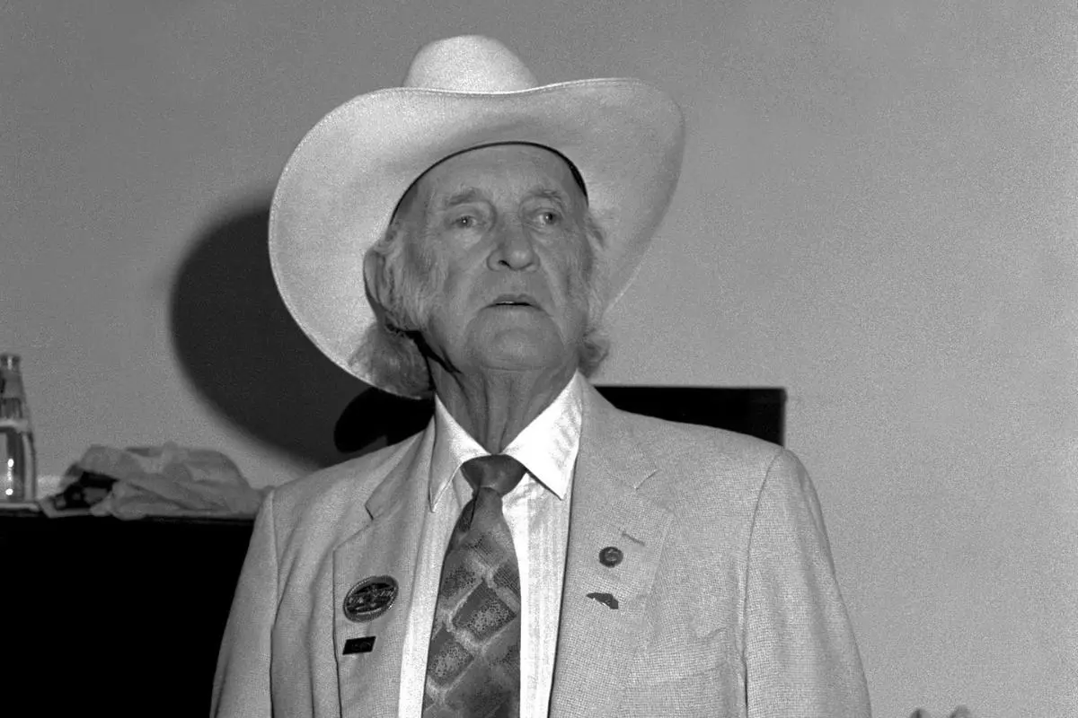 Bill Monroe in a light-colored suit and cowboy hat stands indoors, gazing off to the side under soft lighting.