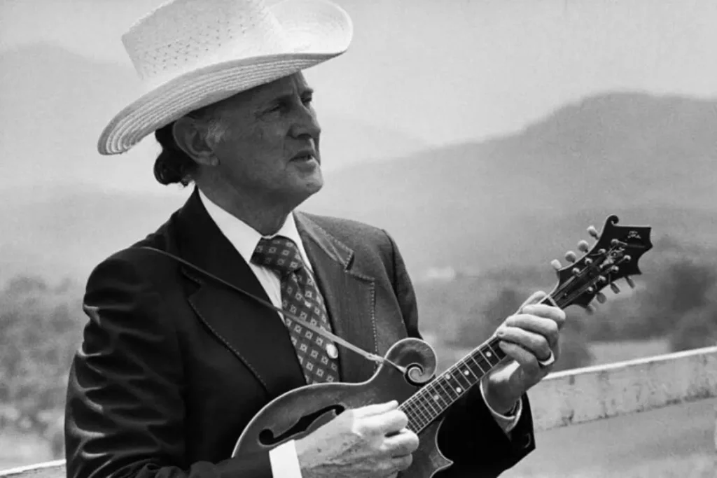 Bill Monroe in a suit and wide-brimmed hat plays a mandolin outdoors, looking focused against a mountainous backdrop.