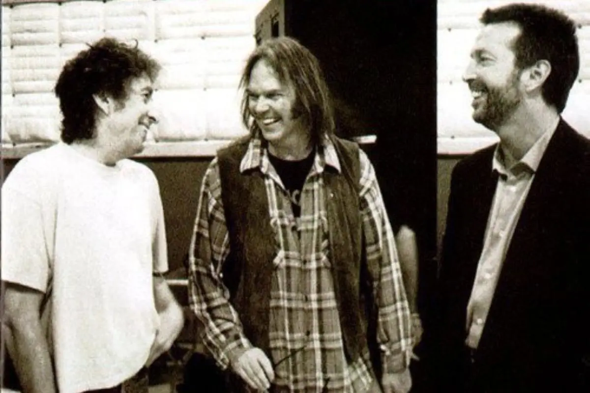 Bob Dylan, Neil Young, and Eric Clapton stand together in a candid backstage moment.
