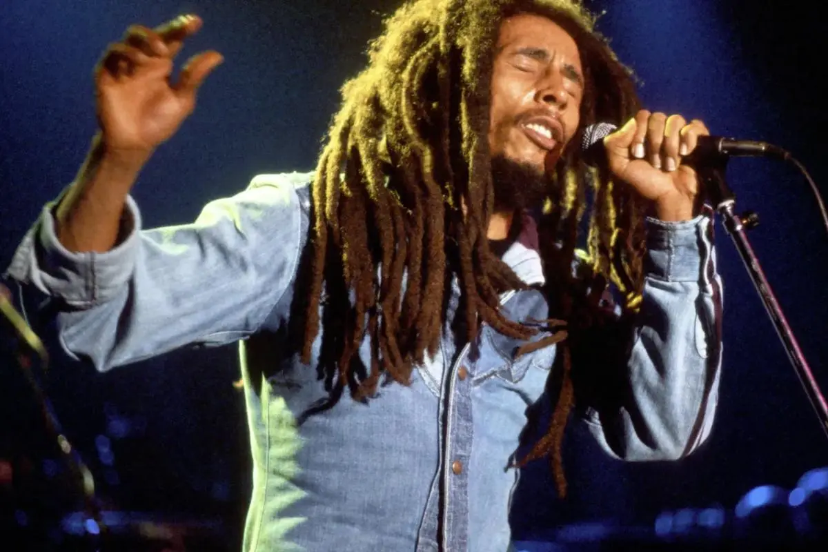 Bob Marley with long dreadlocks sings passionately into a microphone onstage, eyes closed and one hand raised.