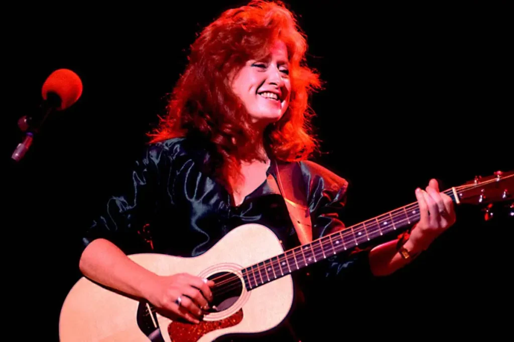 Bonnie Raitt with red hair plays an acoustic guitar onstage under warm red lighting.