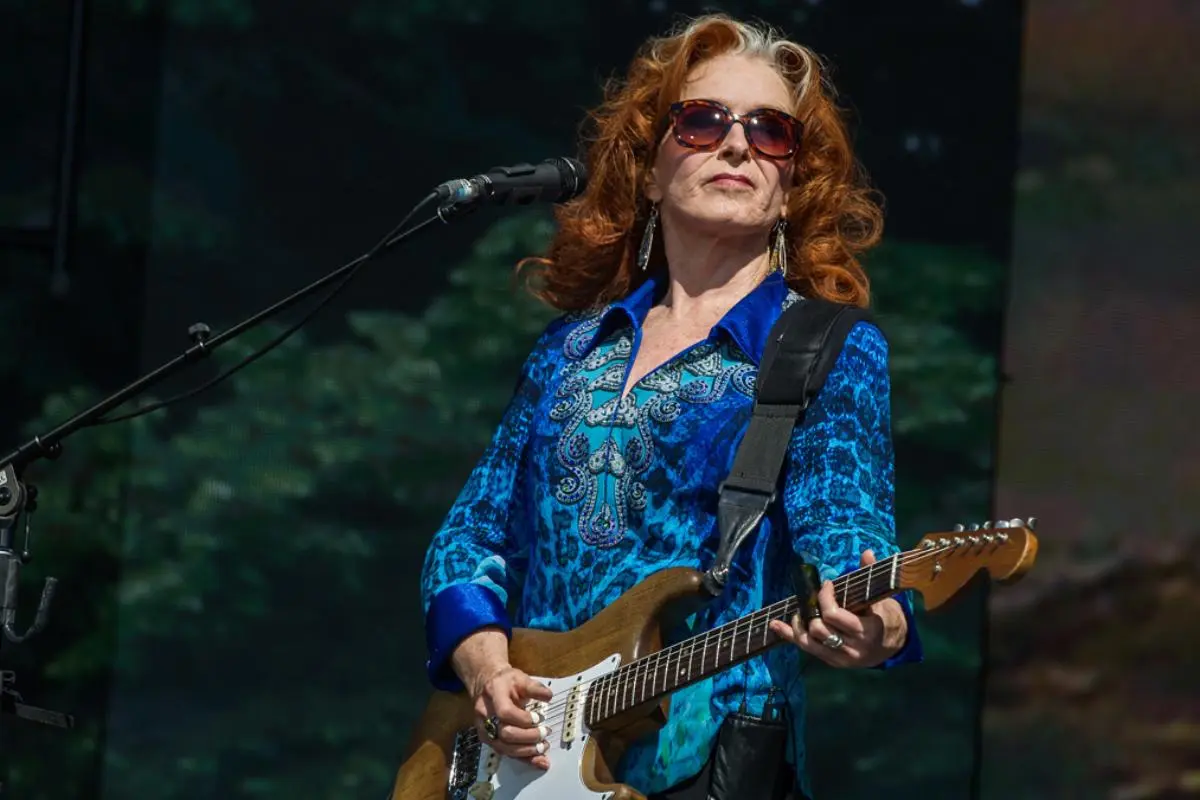 Bonnie Raitt with red hair and sunglasses performs on an outdoor stage, wearing a blue patterned shirt and playing an electric guitar.