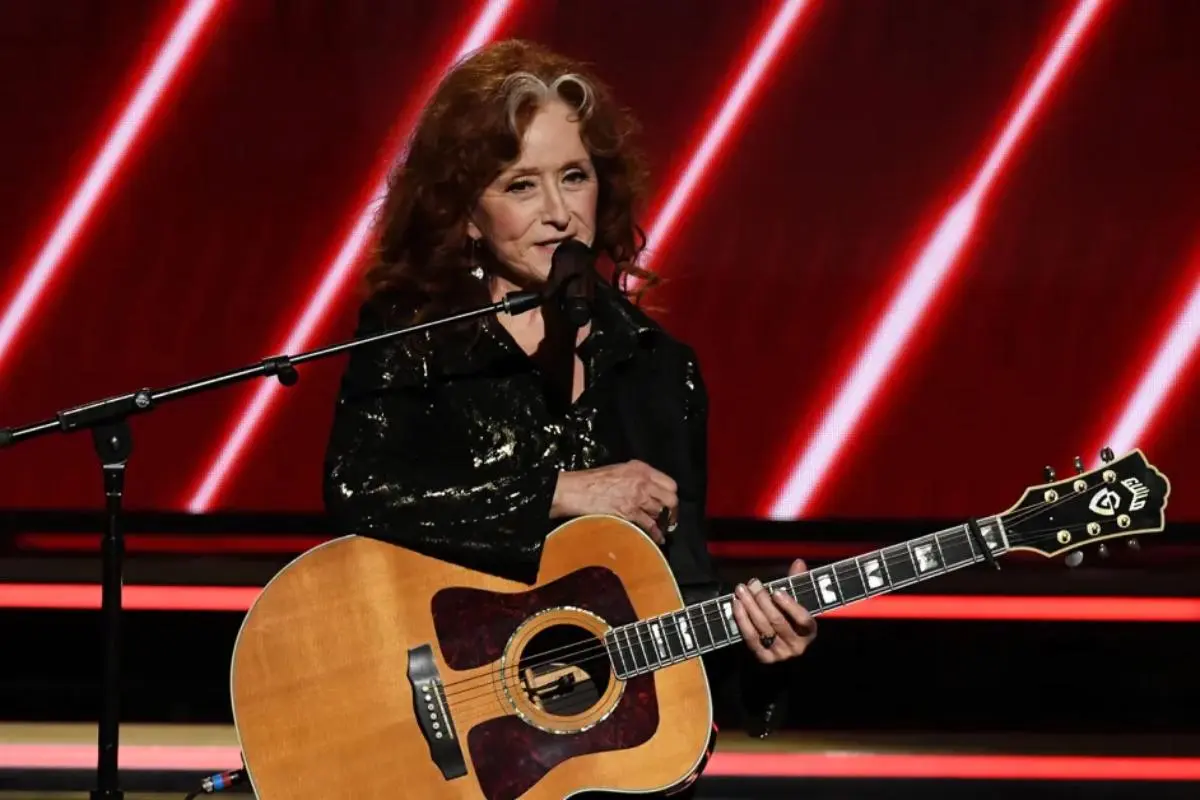 Bonnie Raitt with red hair stands at a microphone holding an acoustic guitar, lit by red diagonal stage lights behind her.