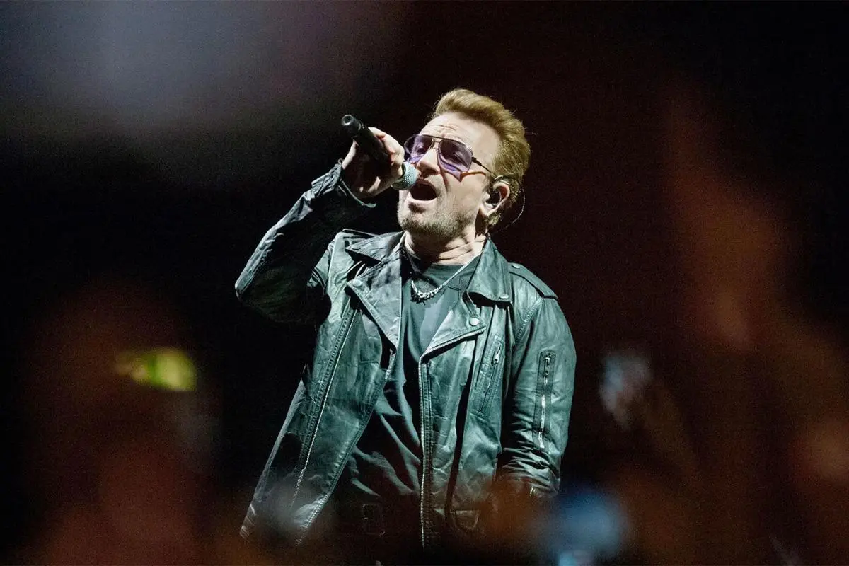Bono performing on stage, singing into a handheld microphone while wearing a black leather jacket and tinted glasses under dramatic concert lighting.
