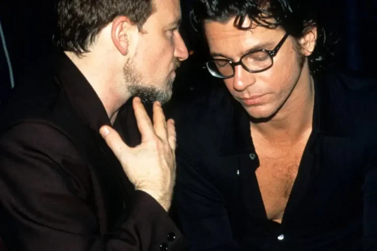 Bono and Michael Hutchence standing closely together backstage, with Bono leaning in as if speaking quietly while Michael Hutchence looks down thoughtfully, both wearing dark clothing.