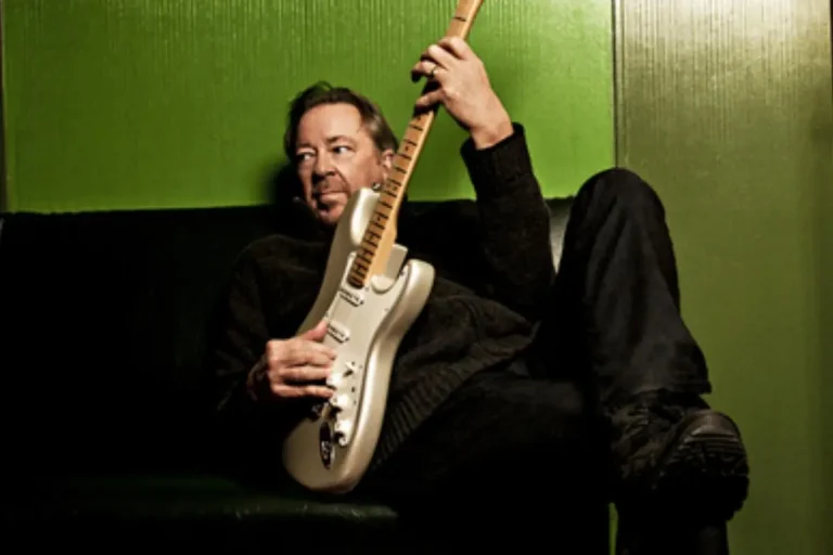 Boz Scaggs reclines on a couch holding an electric guitar, posed casually against a bright green wall.