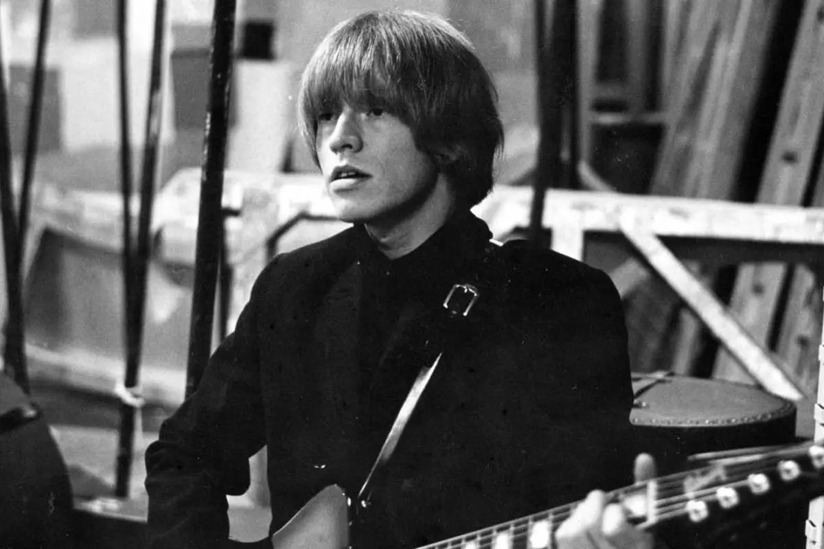 Brian Jones sitting with a guitar, wearing a dark jacket.