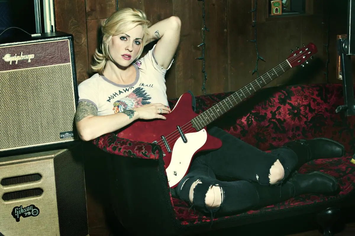 Brody Dalle reclining on a couch holding a red electric guitar beside an amplifier.