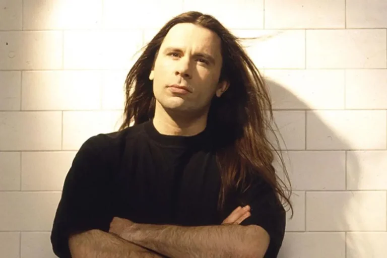 Portrait of Bruce Dickinson with long hair, arms crossed, standing against a white brick wall.