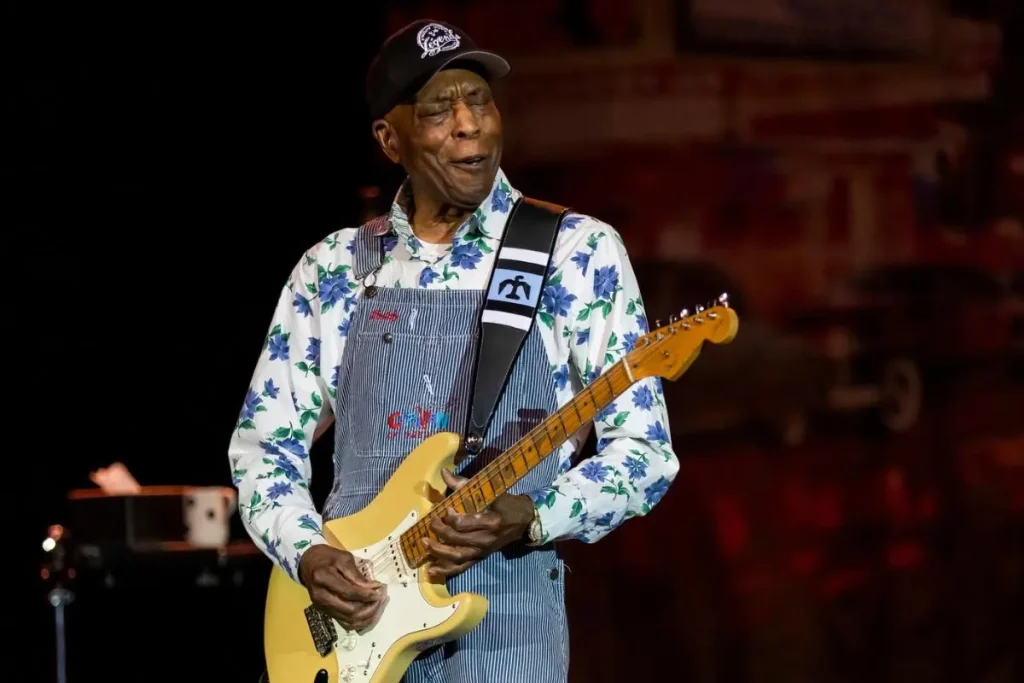 Buddy Guy guitarist performs onstage wearing denim overalls and a floral shirt, playing an electric guitar with an expressive look on his face.