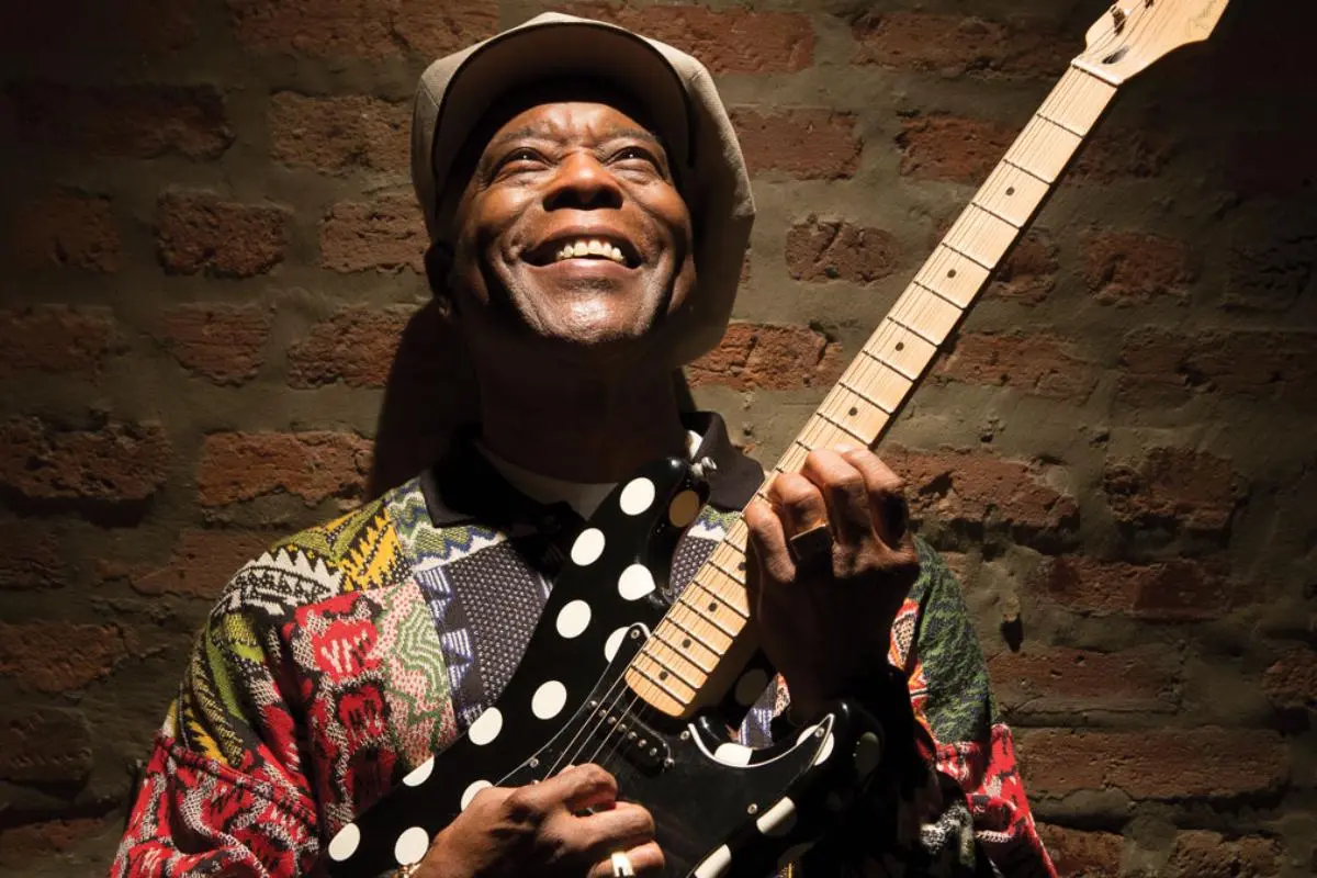 Buddy Guy holds a polka-dot electric guitar while standing against a brick wall, illuminated warmly from above.