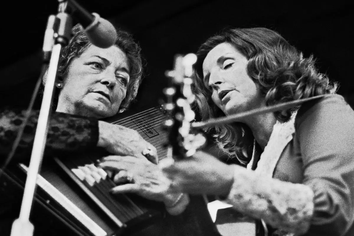 Maybelle Carter plays an autoharp beside a younger woman playing guitar, both leaning in closely and performing together onstage