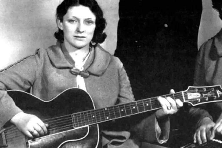 Maybelle Carter sits holding an acoustic guitar in an early black-and-white portrait.