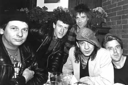 Black-and-white candid photo of a group of musicians seated closely together at a table, capturing a relaxed moment of camaraderie and creative community.