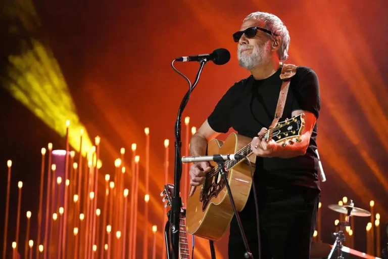 Cat Stevens performing on stage, playing an acoustic guitar under warm stage lights.