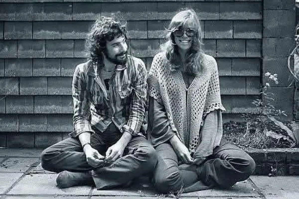 Cat Stevens and Carly Simon sitting outdoors against a wooden wall, smiling casually.