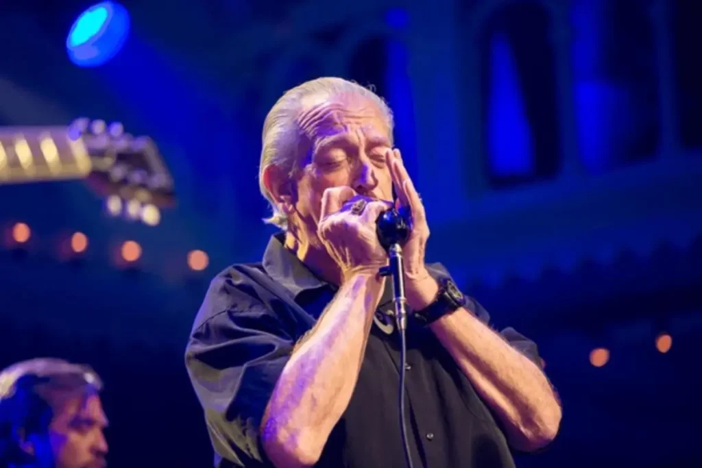 Charlie Musselwhite playing harmonica on stage under blue lighting.