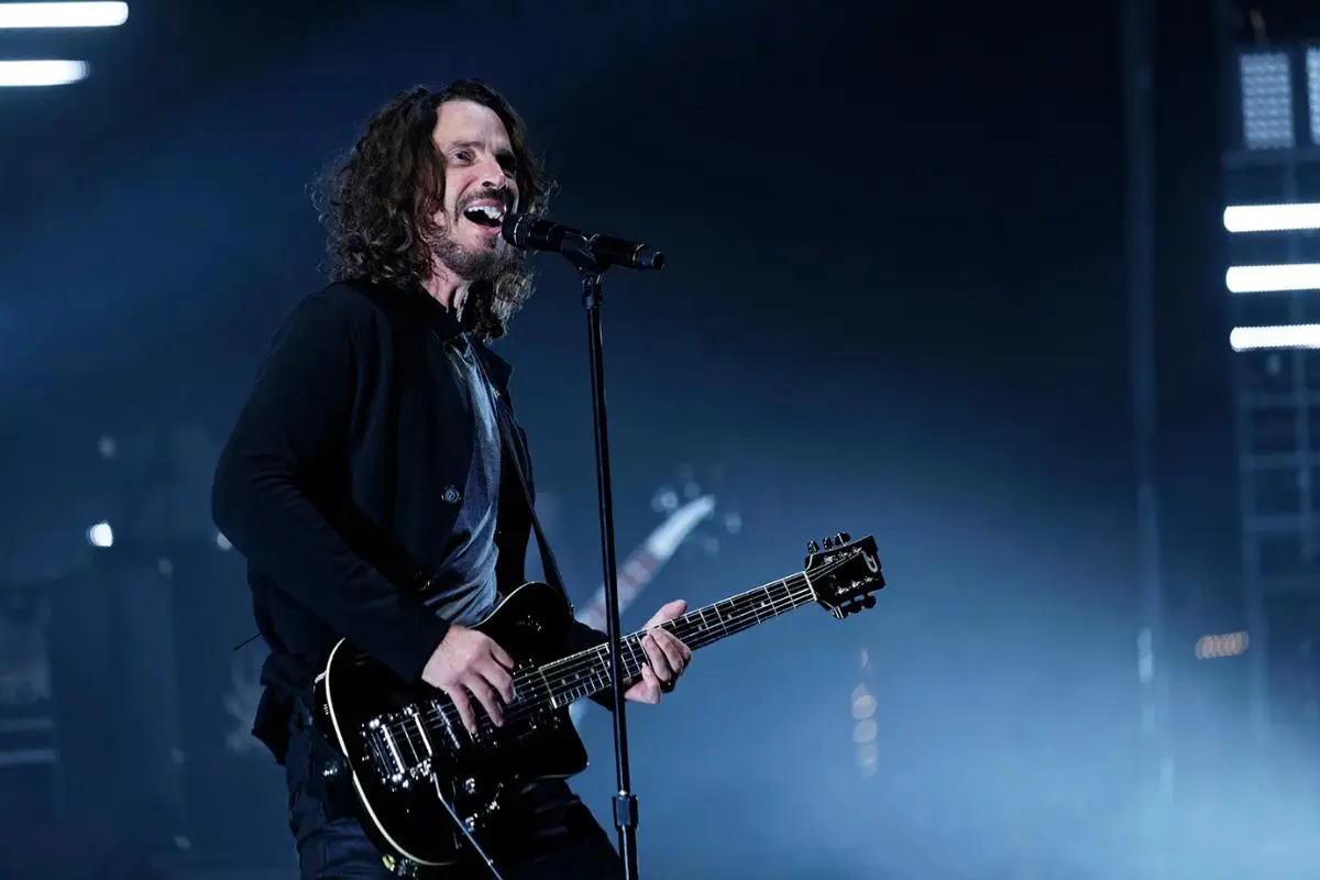 Chris Cornell performs onstage while playing an electric guitar, leaning into the microphone under cool blue lighting.