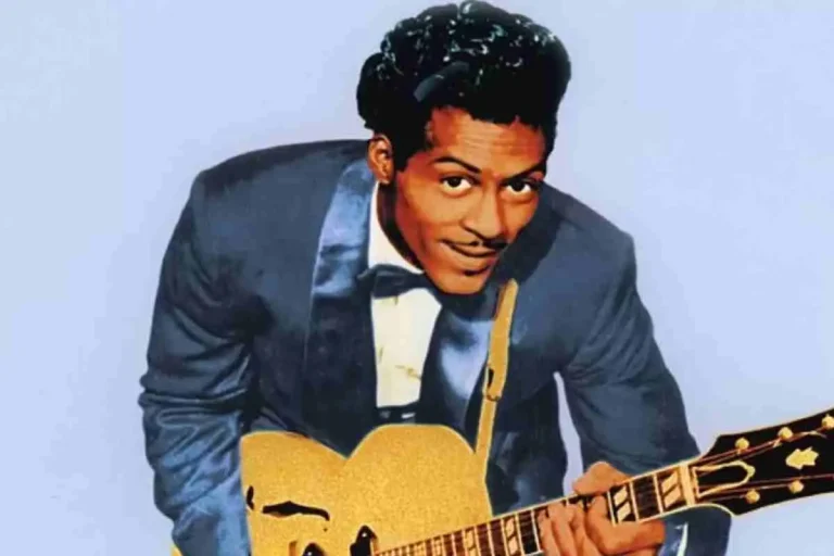 Chuck Berry leaning forward while playing a gold electric guitar, wearing a blue suit jacket and bow tie against a light background.