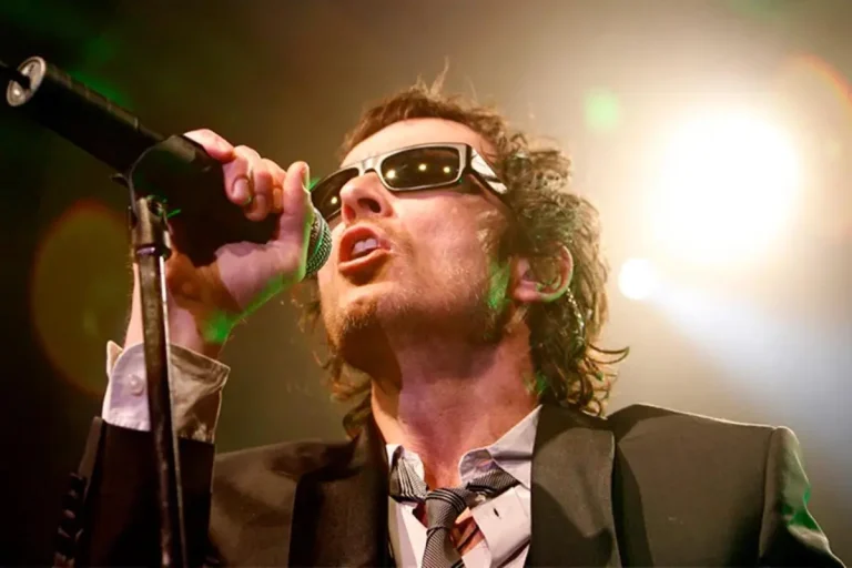 Chuck Negron onstage singing into a microphone and playing a pale semi-hollow electric guitar.