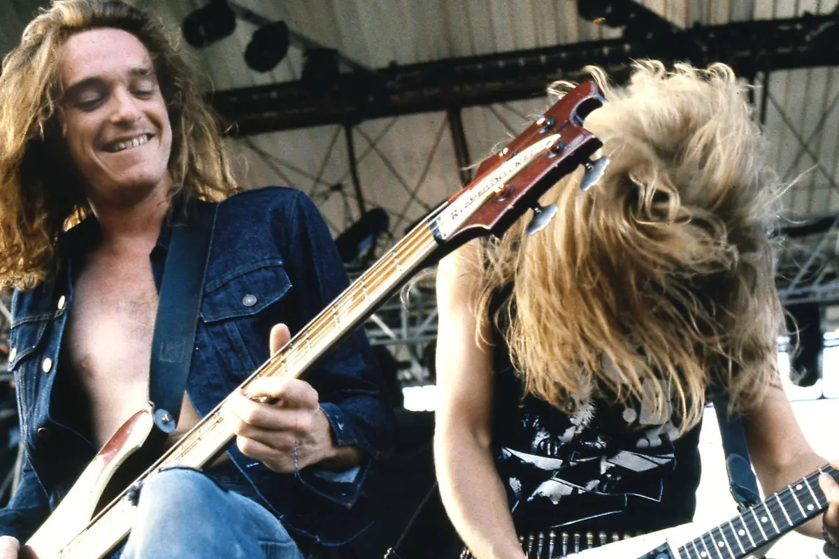 Cliff Burton performing on stage, one smiling while playing bass as the other headbangs on guitar, capturing the raw energy of a live metal performance.