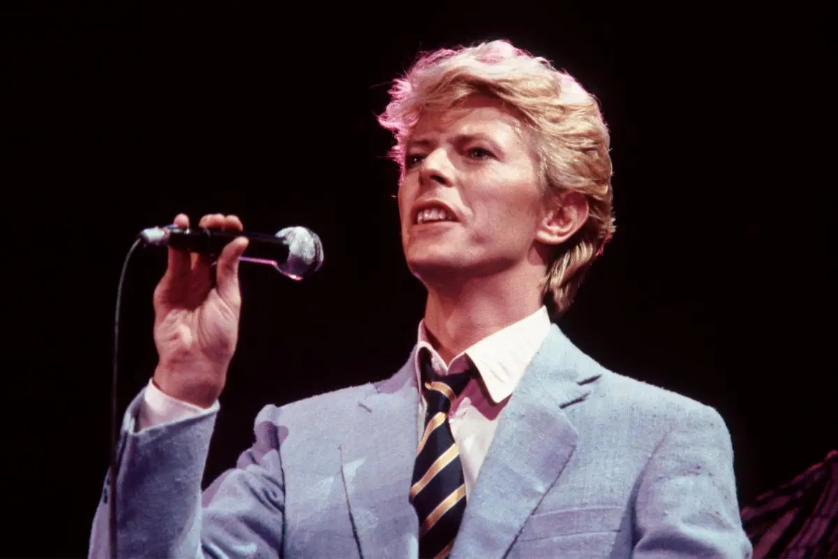 David Bowie performs onstage in a light-blue suit, holding a microphone under stage lights.