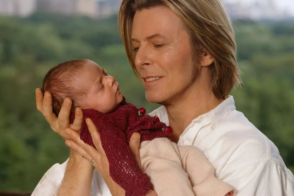 David Bowie holding a baby while gazing at them outdoors, conveying tenderness, care, and a strong parent-child bond.
