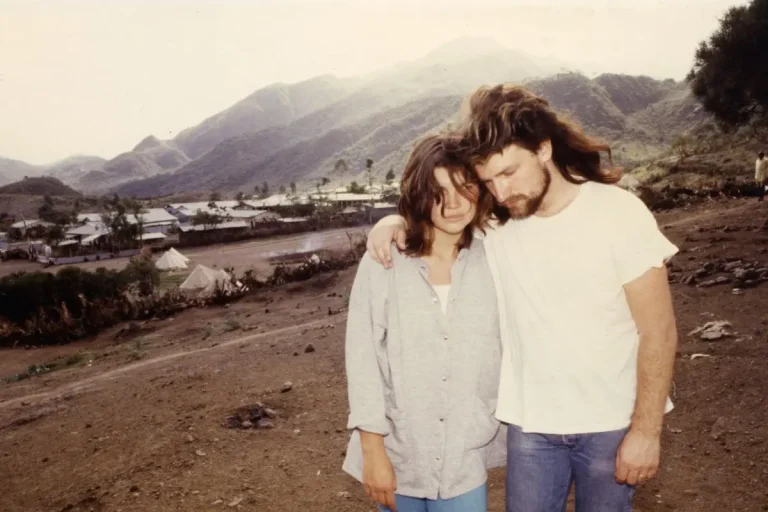 Bono and Ali Hewson stands on a dusty hillside with mountains and a rural settlement behind them, leaning into each other with somber expressions.