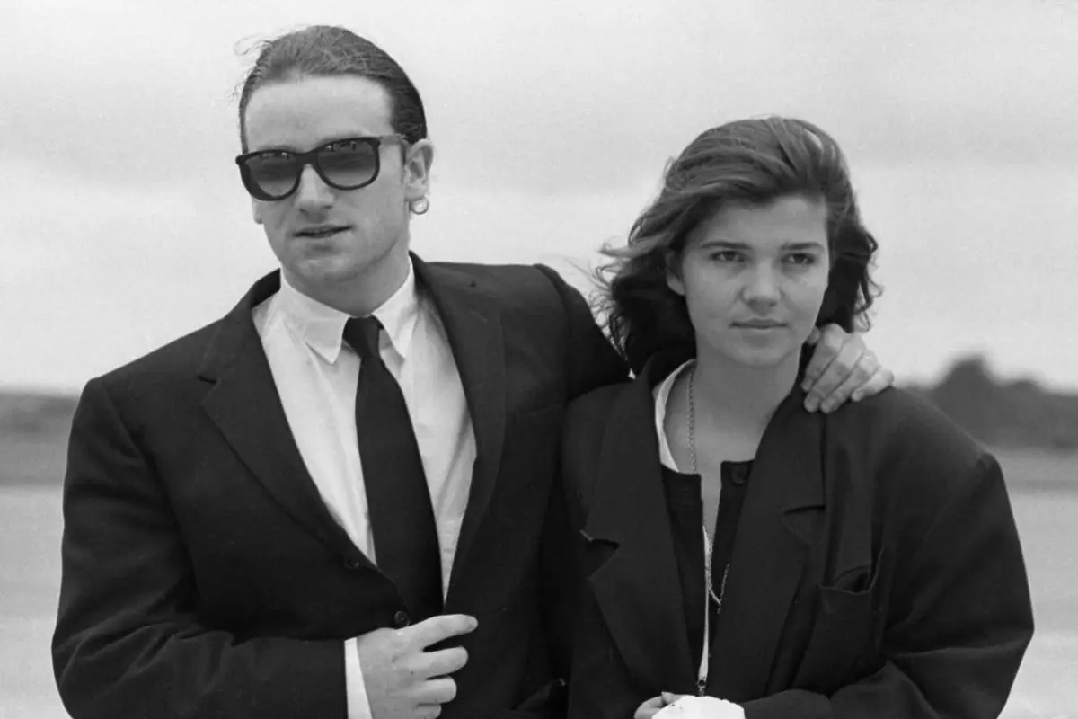 Bono Hewson in a suit and dark sunglasses stands outdoors with his arm around Ali Hewson dressed in a blazer, both looking ahead with calm expressions.