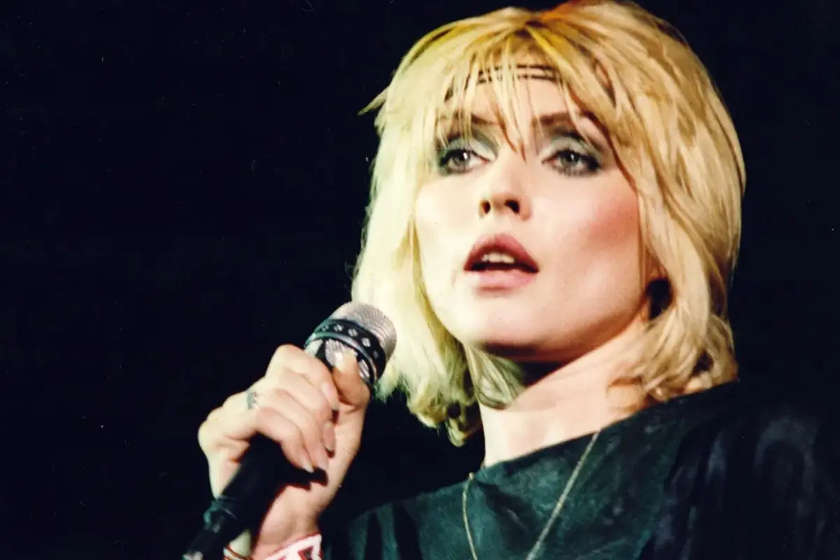 Debbie Harry holding a microphone on stage, captured mid-song with an intense, focused expression under dramatic concert lighting.