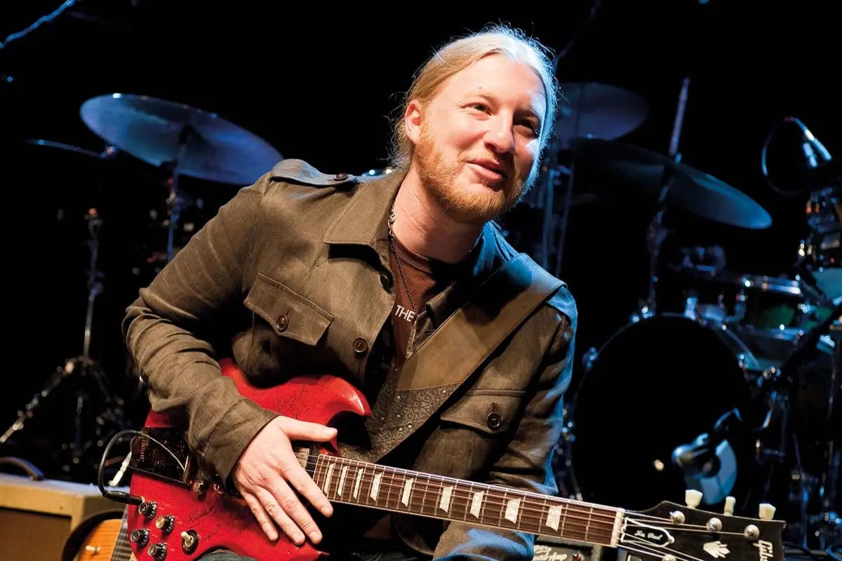 Derek Trucks seated on stage holding an electric guitar and smiling, suggesting a relaxed moment of connection with the audience between songs.