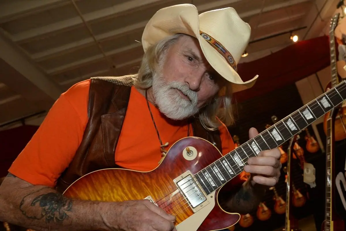 Dickey Betts wearing a cowboy hat while playing guitar indoors.