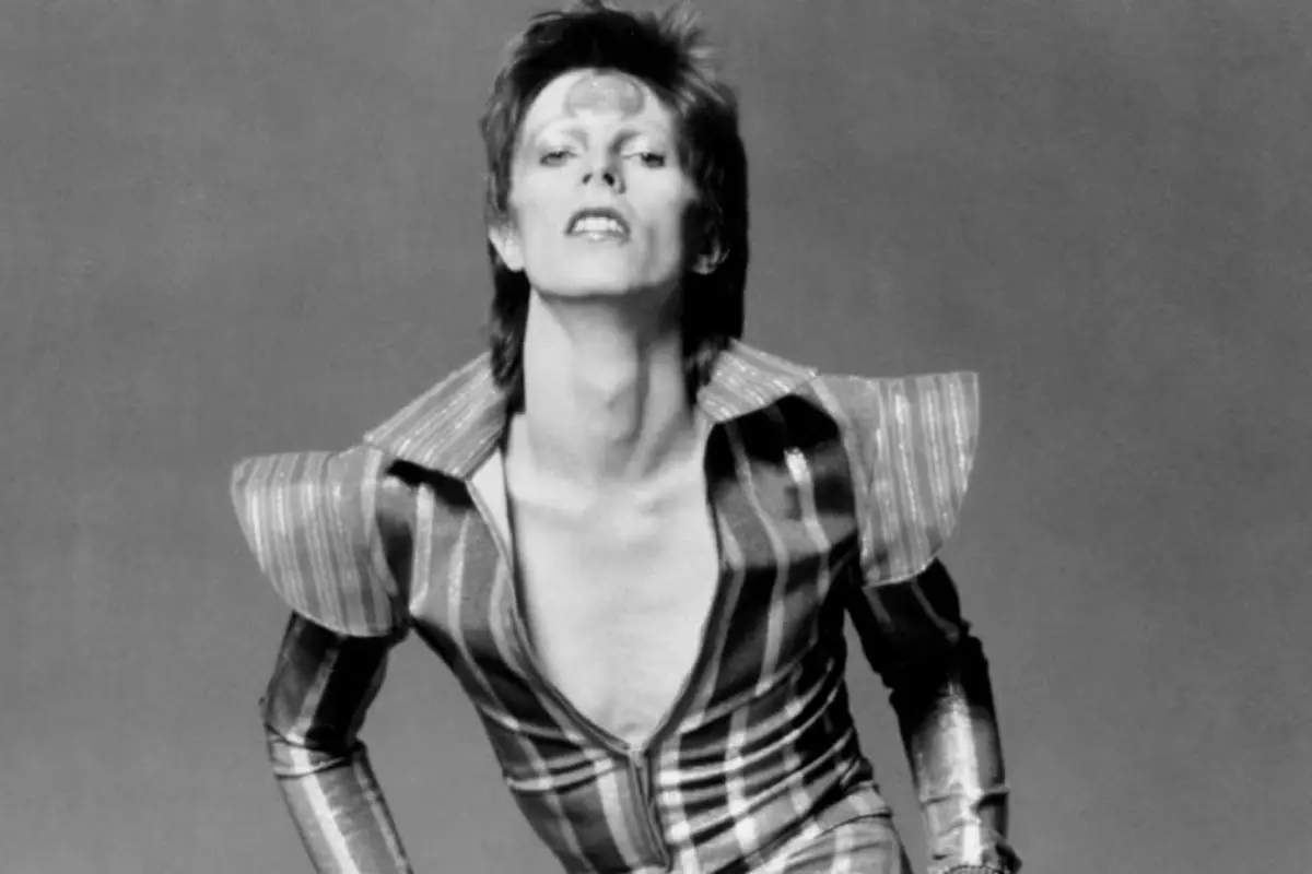  David Bowie in a striped, futuristic jumpsuit leans forward confidently, wearing dramatic stage makeup and styled hair against a plain backdrop.