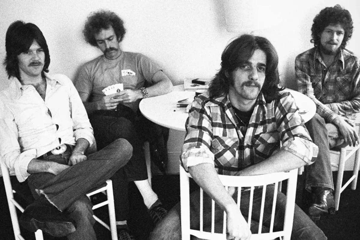 Eagle's band sit around a small table in a casual room, relaxed and informal, captured in a black-and-white photograph.