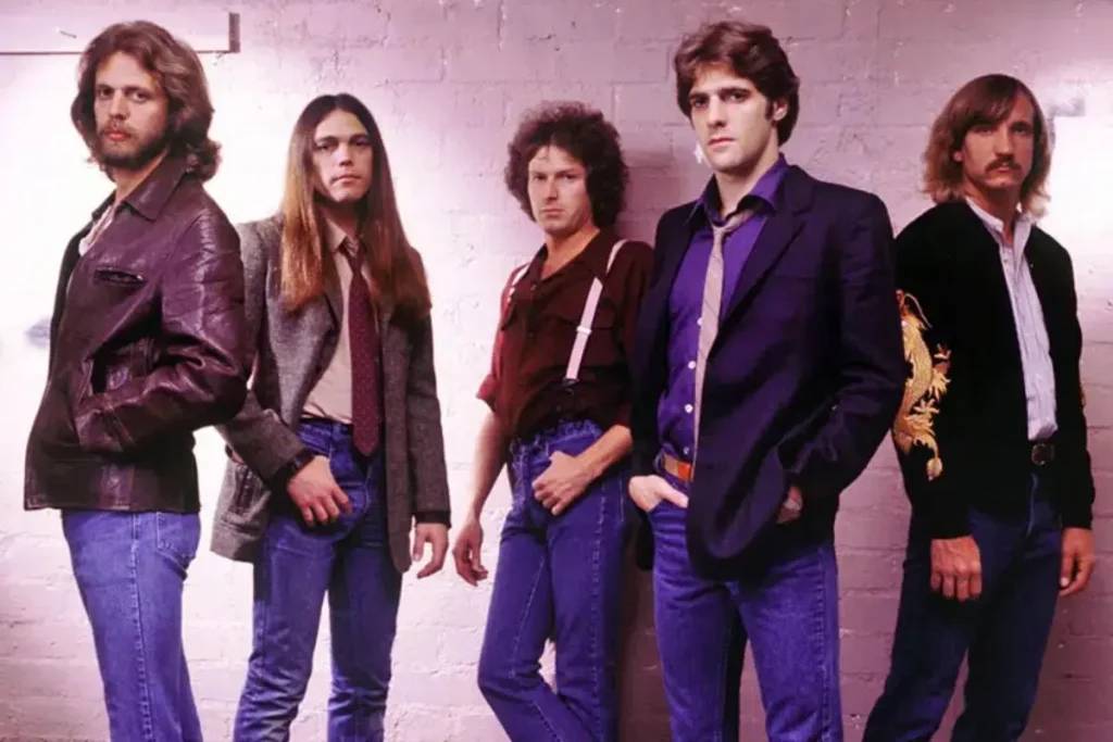Eagle's band stand against a wall wearing denim and jackets, styled in a late-1970s rock band aesthetic.